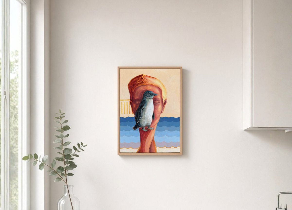 A framed art print featuring a stylized illustration of a penguin on a camel hump, hanging on a light beige wall above a white kitchen counter with a glass vase of eucalyptus.