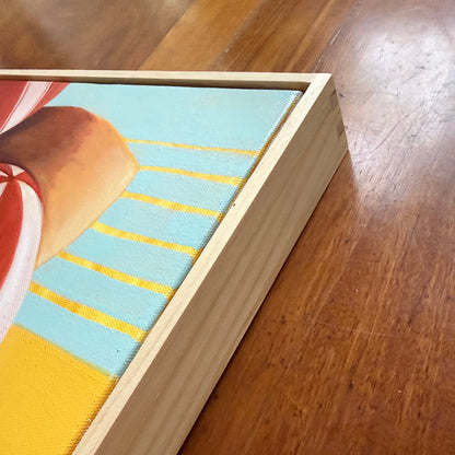 A close-up of a framed canvas painting resting on a polished wooden floor. The artwork features a light blue background with yellow diagonal stripes and a warm tan shape, housed in a natural light wood frame.