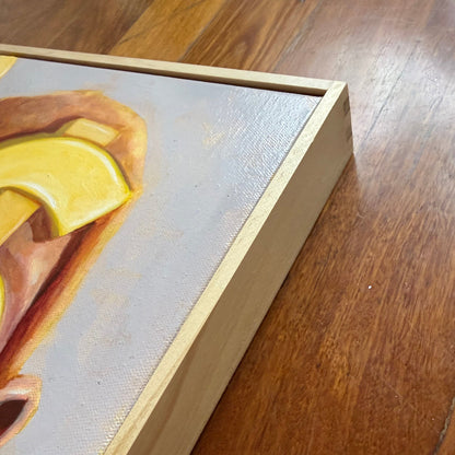 A close-up of a framed painting on a wooden floor, showing part of the artwork with yellow and orange tones on a light gray canvas, along with the natural wood frame.