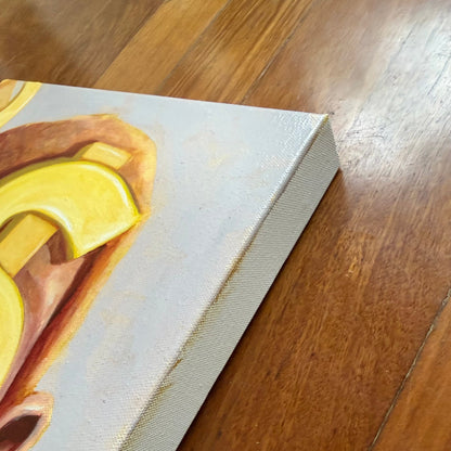 A close-up of a canvas painting resting on a polished wooden floor, showing the textured canvas edge and a partial abstract yellow and brown design on a light gray background.