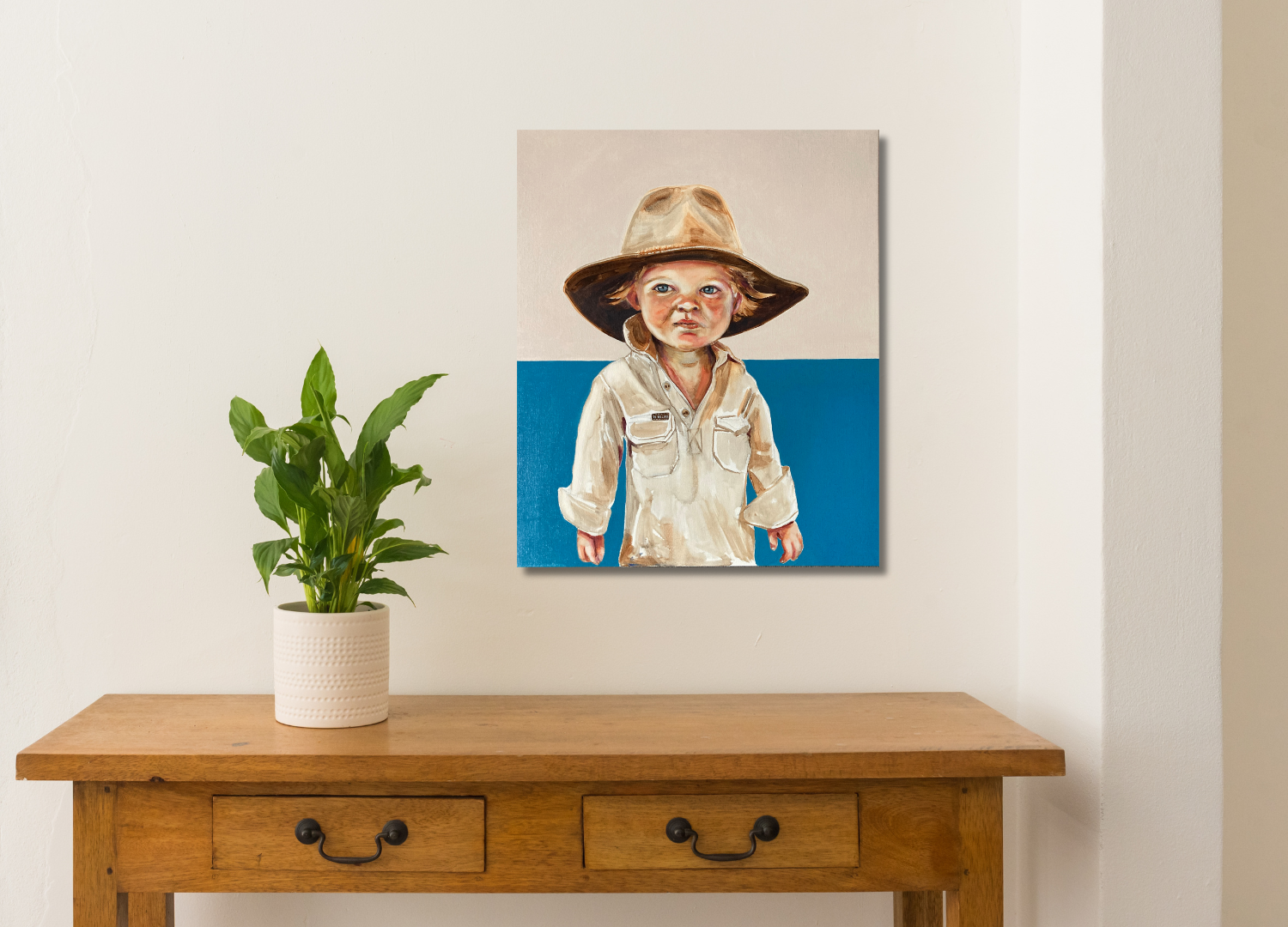 A hand-painted portrait of a child wearing a hat, set against a blue background, displayed on a wooden table with plant