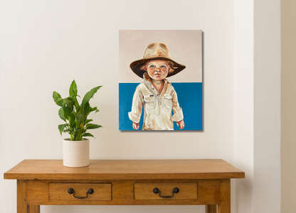 A hand-painted portrait of a child wearing a hat, set against a blue background, displayed on a wooden table with plant